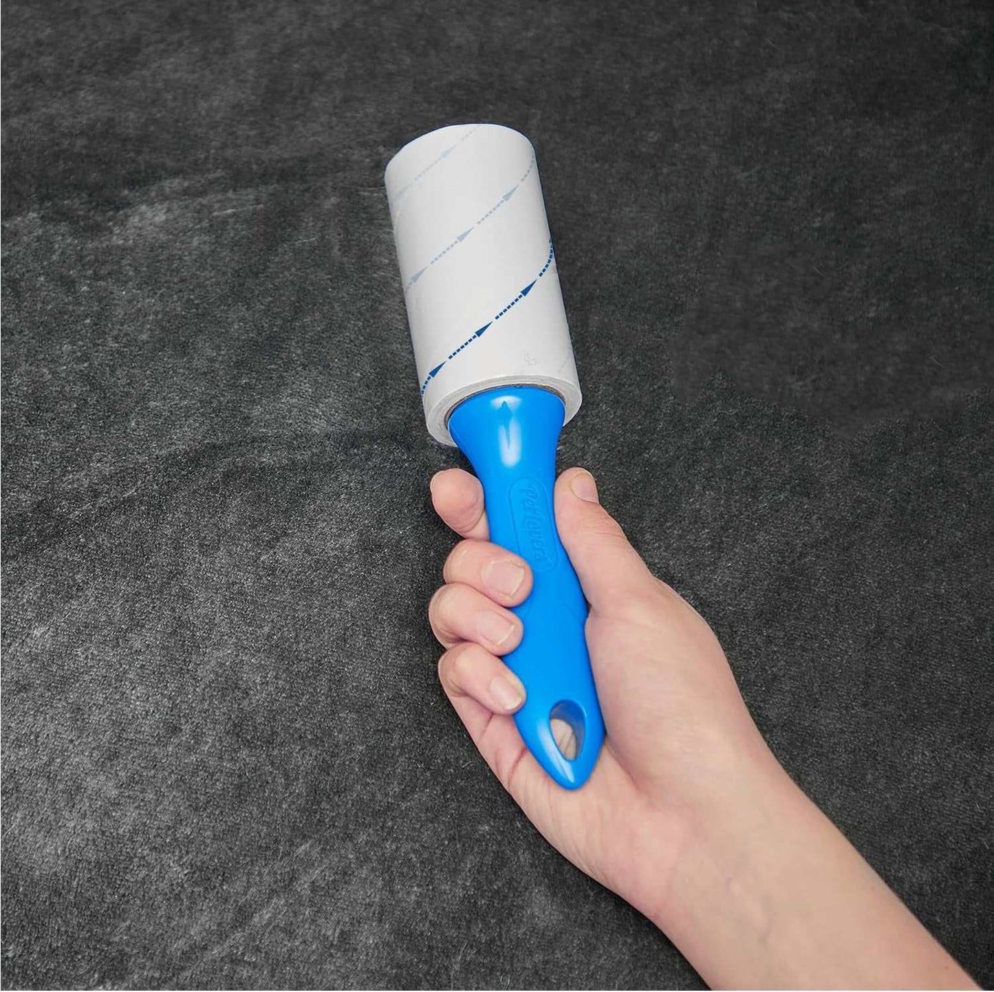 Hand holding a blue-handled pet hair removal lint roller with extra sticky sheets on a dark surface