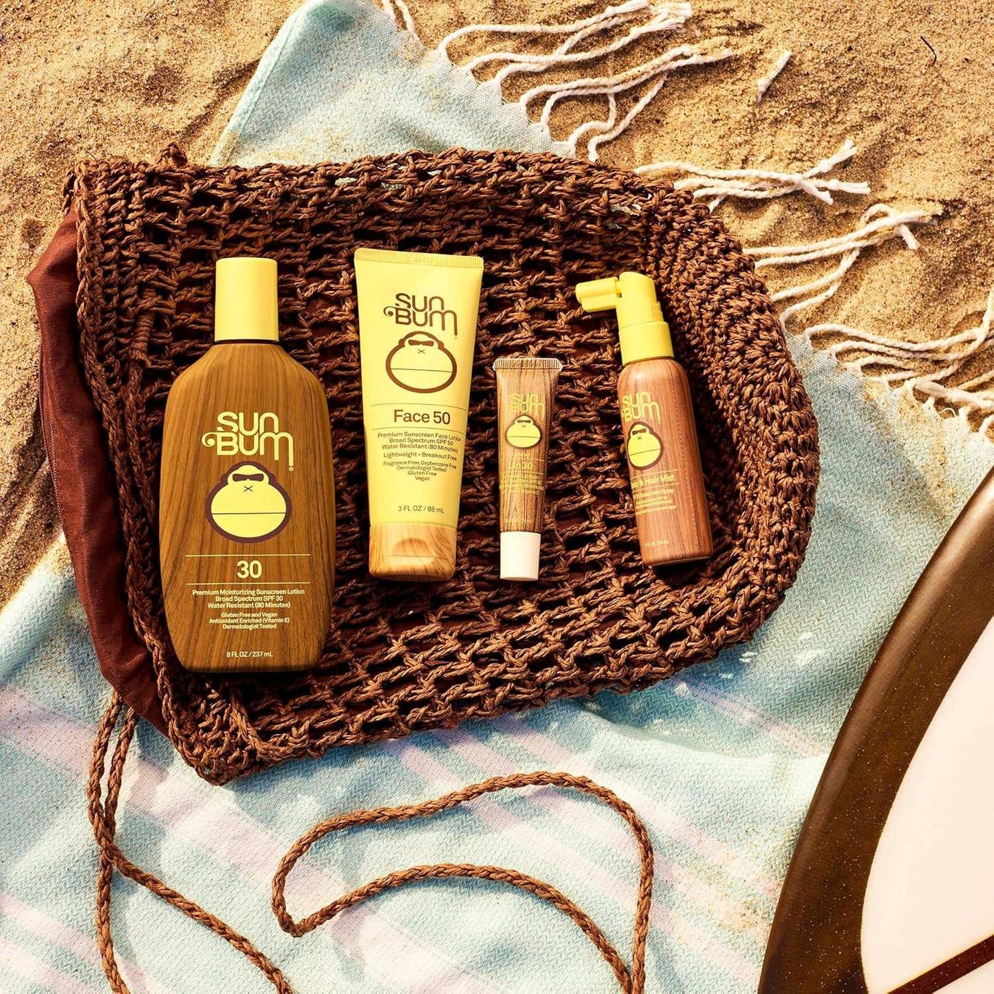Sun Bum sunscreen products including SPF 30 lotion, SPF 50 face sunscreen, moisturizing lip balm with SPF, and spray sunscreen on woven mat and beach towel