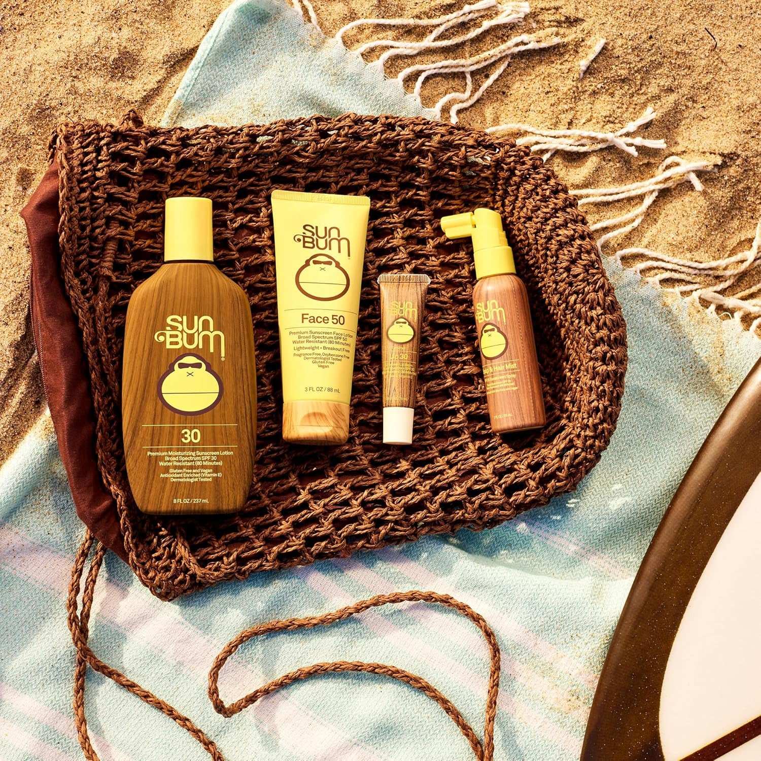 Sun Bum sunscreen products including SPF 30 lotion, SPF 50 face sunscreen, moisturizing lip balm with SPF, and spray sunscreen on woven mat and beach towel