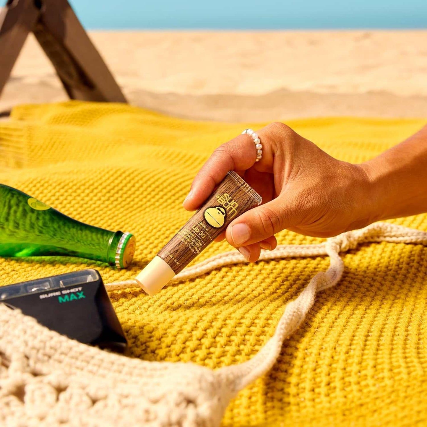 Hand holding reef-safe moisturizing sunscreen lip balm tube on yellow textured cloth at the beach